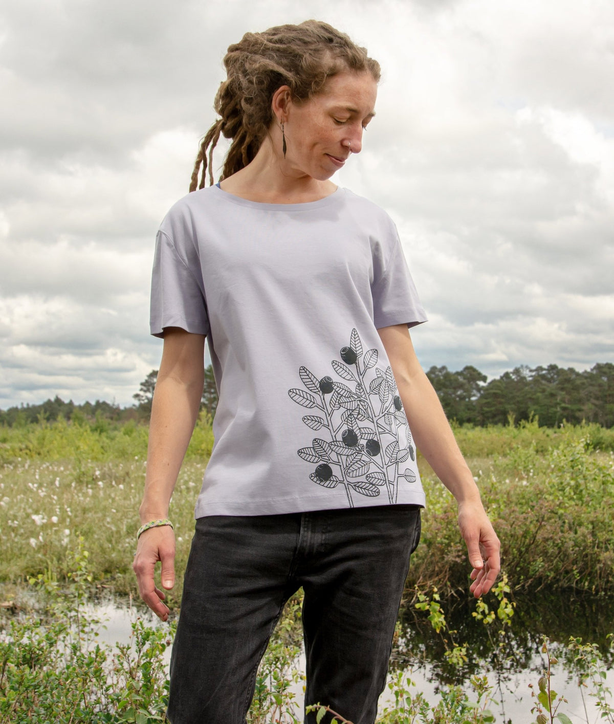 Blaubeere Tshirt für Damen aus dickerem Bio-Stoff (180g/m²). Ein herbstliches Outdoor-Lifestyle-Bild zeigt eine Frau in Ganzkörperansicht in der Natur mit dem handbedruckten lavendelfarbenen Oberteil.