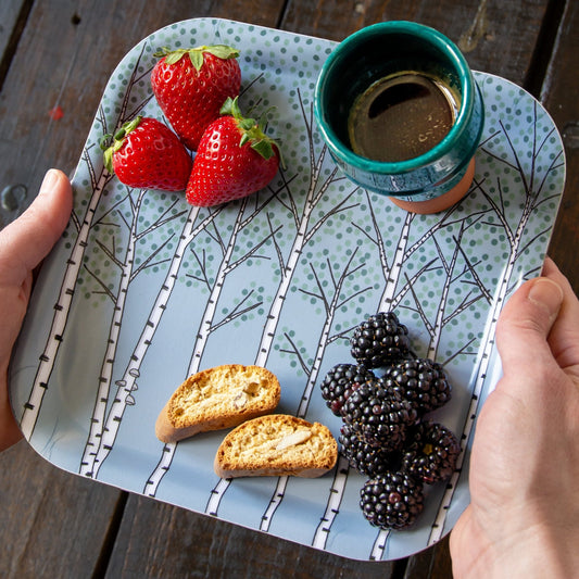 Kleines quadratisches Holztablett im skandinavischen Design beim Servieren. Eine Hand trägt das hellblaue Tablett mit Birkenwald-Motiv für die schwedische Fika-Kaffeepause.