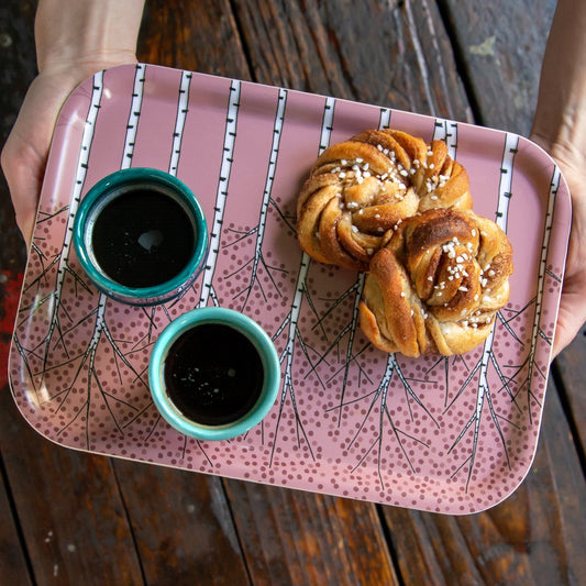 Schwedisches Design Frühstücksblett aus holz beim Servieren in der Küche. Eine Person trägt das altrosa Serviertablett mit Birken-Muster, beladen mit einer Kaffeetasse und einer frischen Zimtschnecke für die Fika-Pause.