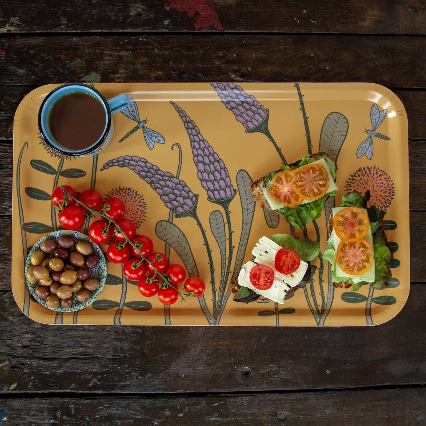 Schwedisches Design Holztablett reichlich gedeckt für das Frühstück. Großes rechteckiges Serviertablett in Gelb mit Blumenwiese-Motiv und libellen, beladen mit teetassen, belegten Broten, Oliven und Tomaten.