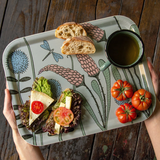Schwedisches Design Holztablett beim Servieren von Frühstück im Garten. Eine Person trägt das salbeigrüne Serviertablett mit Blumenwiese-Motiv, beladen mit frischem Tee, belegten Broten und Tomaten.