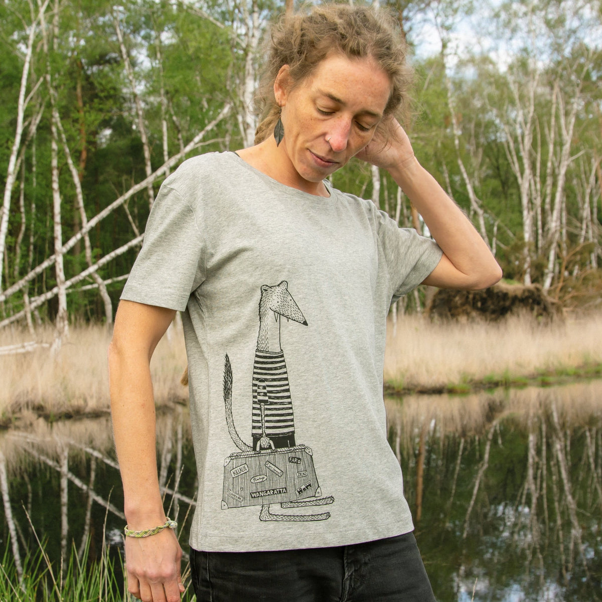 Wiesel T-Shirt Kollektion aus weichem Bio-Stoff für den Urlaub. Eine Frau präsentiert das handbedruckte Kurzarm-Shirt mit dem gut sichtbaren Koffer-Frontprint in einer klaren Studio- oder Natur-Ansicht.