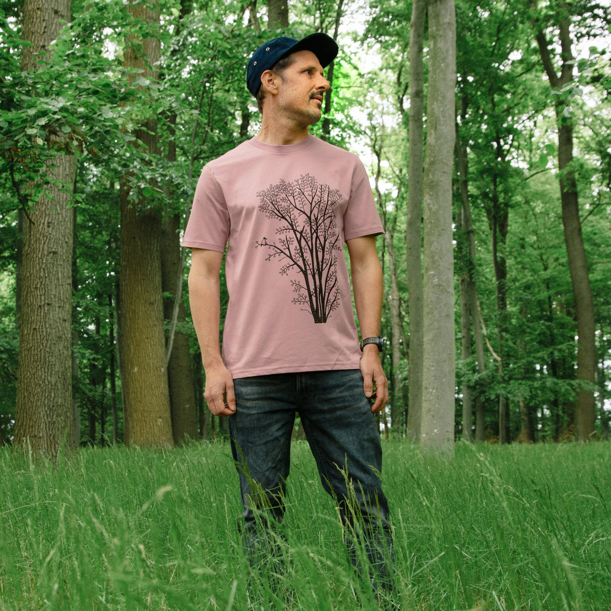 Baum T-Shirt für Männer im lockeren Unisex-Schnitt. Ein Mann trägt das handbedruckte, lila-graue Naturmode-Shirt aus Bio-Baumwolle in einer stehenden Outdoor-Ansicht.
