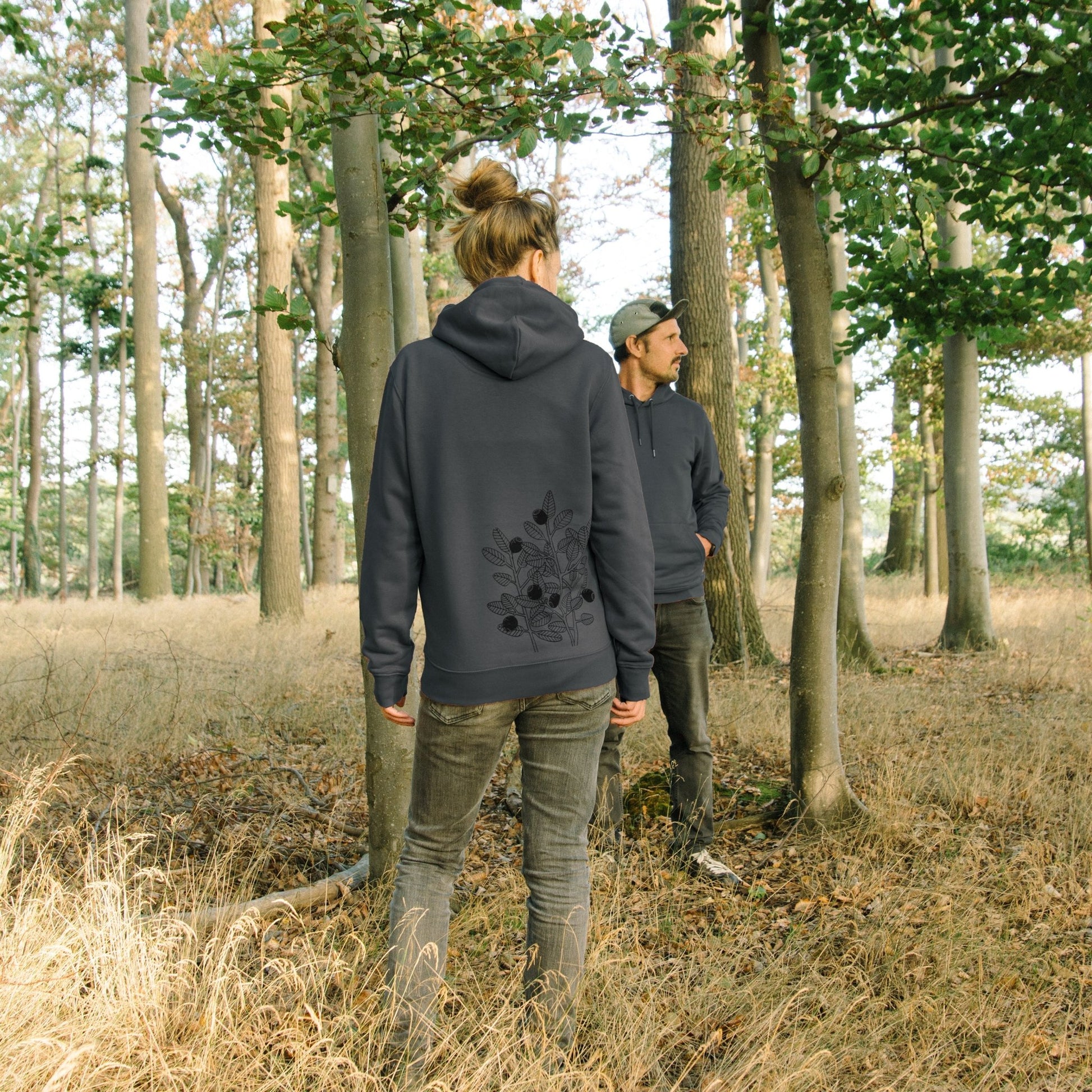Blaubeere Hoodie aus Bio-Baumwolle für Damen und Herren. Ein herbstliches Outdoor-Lifestyle-Bild im Wald zeigt ein Pärchen in den gemütlichen, dunkelgrauen Kapuzenpullis.
