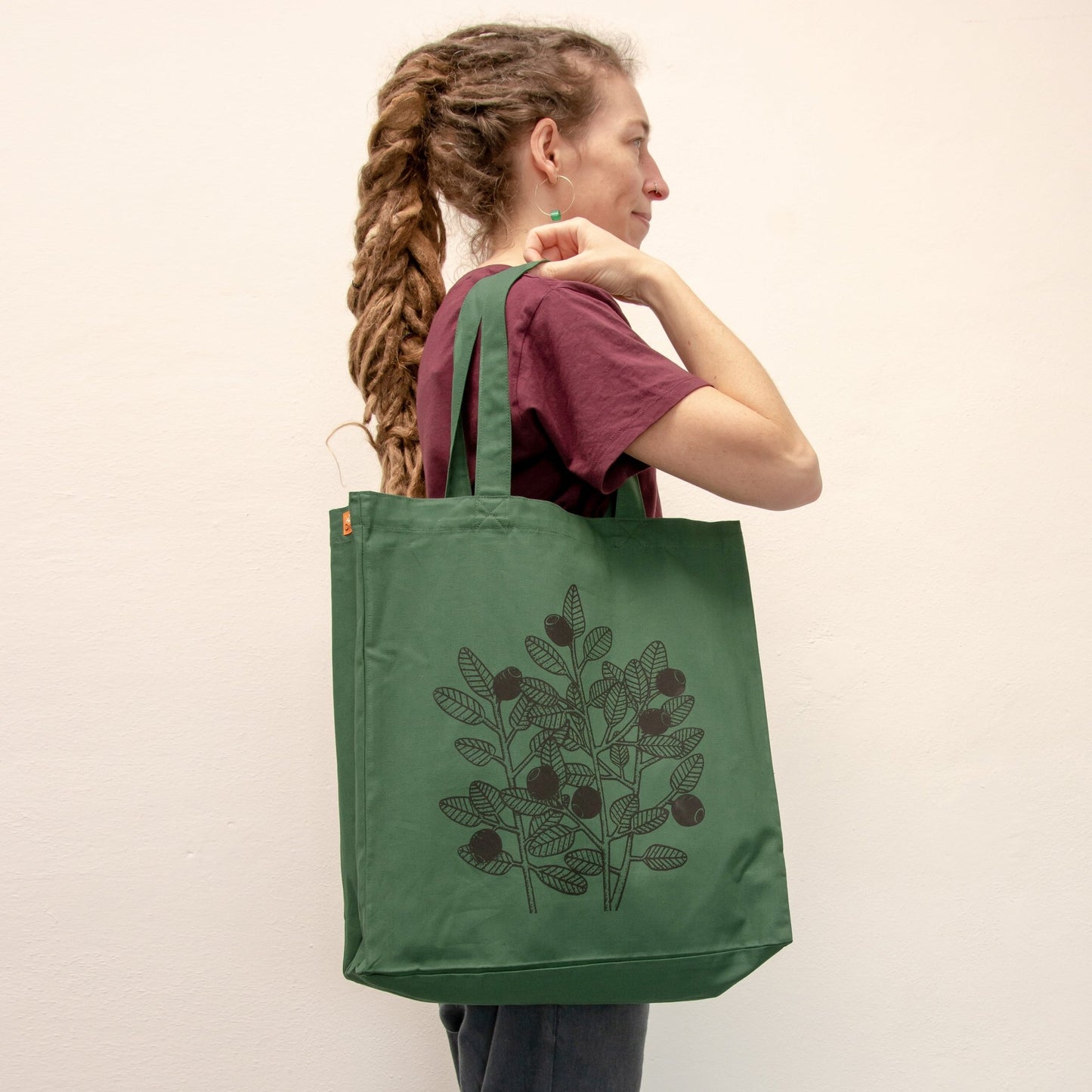 Blaubeere Jutebeutel in Bottle Green getragen von einer Frau. Der waldgrüne Shopper mit Naturmotiv zeigt sich als praktischer Alltagsbegleiter und kreatives Geschenk für Frauen.
