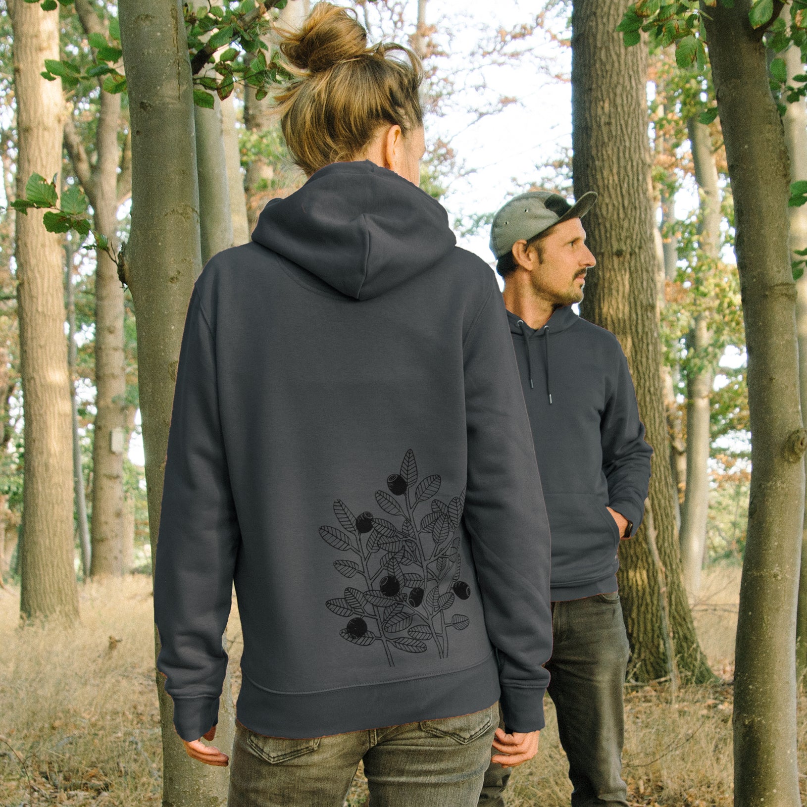 Blaubeere Pullover im Unisex-Schnitt in India Ink Gray. Eine Frau und ein Mann präsentieren gemeinsam den dunkelgrauen Partnerlook-Kapuzenpullover im Wald von vorne und hinten.
