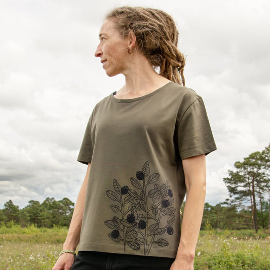 Blaubeere T-Shirt für Damen im lockeren, kurzen Schnitt. Eine Frau steht in der Natur und präsentiert die Vorderansicht des khaki farbenen Bio-Shirts mit dem Beeren-Frontprint an der Seite.
