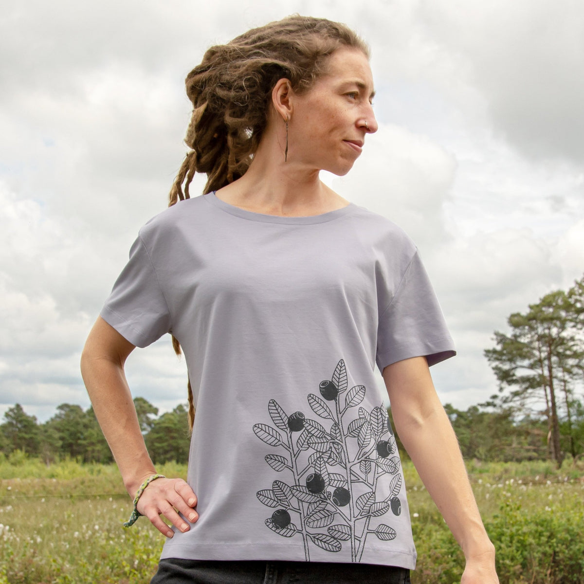 Blaubeere T-Shirt für Damen im lockeren, kurzen Schnitt. Eine Frau steht in der Natur und präsentiert die Vorderansicht des helllila Lavender Bio-Shirts mit dem Beeren-Frontprint an der Seite.
