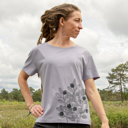 Blaubeere T-Shirt für Damen im lockeren, kurzen Schnitt. Eine Frau steht in der Natur und präsentiert die Vorderansicht des helllila Lavender Bio-Shirts mit dem Beeren-Frontprint an der Seite.
