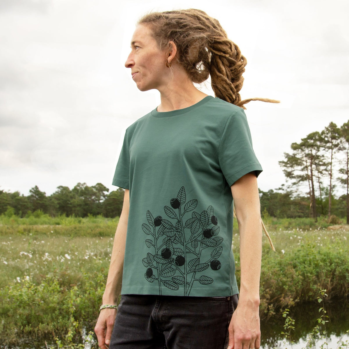 Blaubeere T-Shirt für Damen im lockeren, kurzen Schnitt. Eine Frau steht in der Natur und präsentiert die Vorderansicht des grünen Green Bay Bio-Shirts mit dem Beeren-Frontprint an der Seite.
