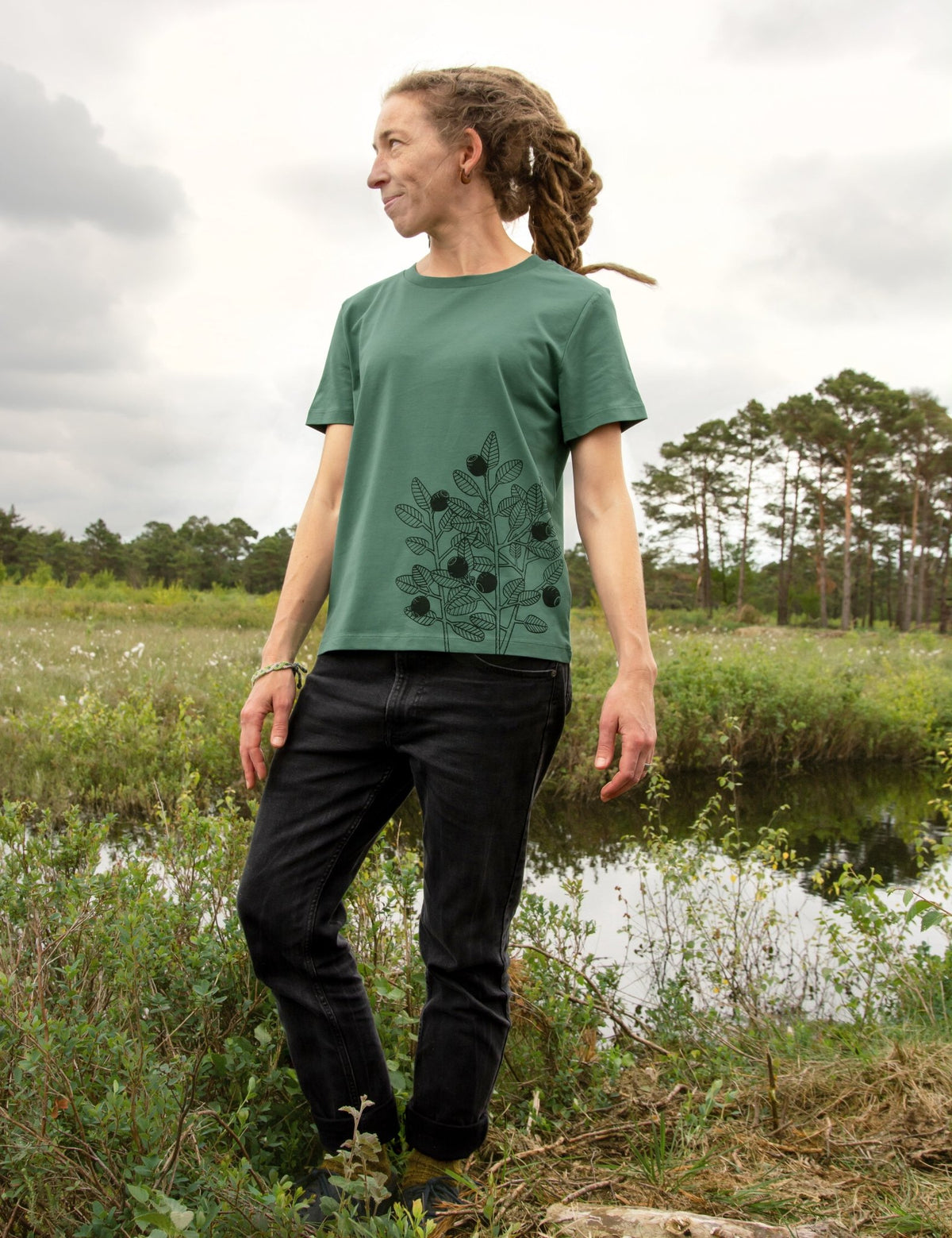 Blaubeere Tshirt für Damen aus dickerem Bio-Stoff (180g/m²). Ein herbstliches Outdoor-Lifestyle-Bild zeigt eine Frau in Ganzkörperansicht in der Natur mit dem handbedruckten Green Bay Oberteil.
