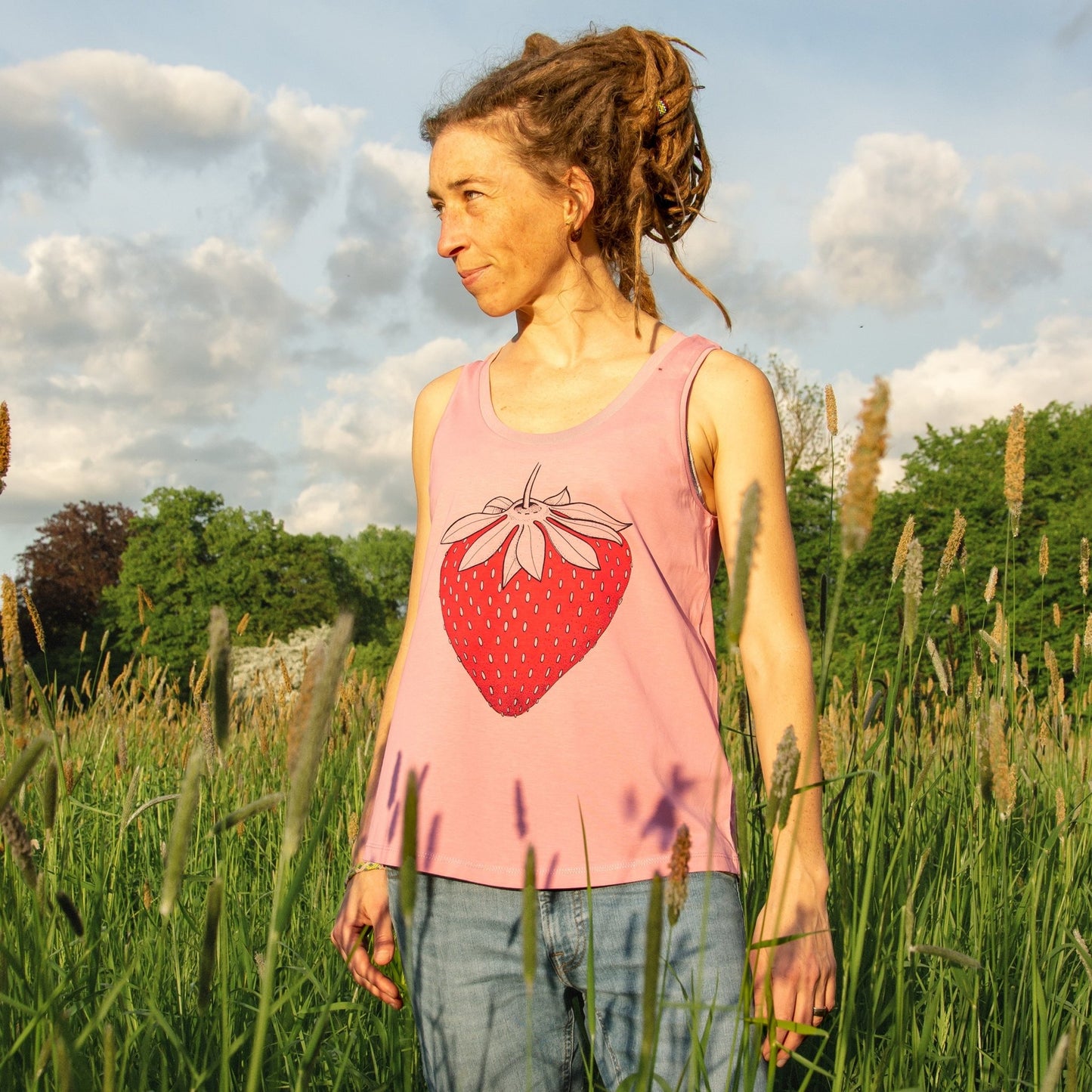 Erdbeer Tanktop Kollektion im bequemen Unisex-Schnitt für Damen. Eine Frau präsentiert das ärmellose Bio-Baumwoll-Shirt mit der großen Erdbeer-Illustration in aufrechter Vorderansicht.
