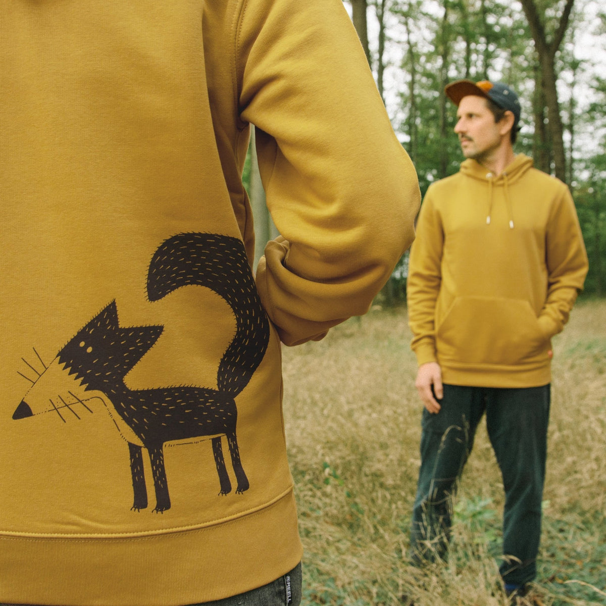 Fuchs Hoodie mit dem detailreichen Franzi-Fuchs-Backprint im Vordergrund. Die Nahaufnahme zeigt die hohe Druckqualität des Siebdrucks, im Hintergrund steht ein Mann im ockerfarbenen Kapuzenpullover.