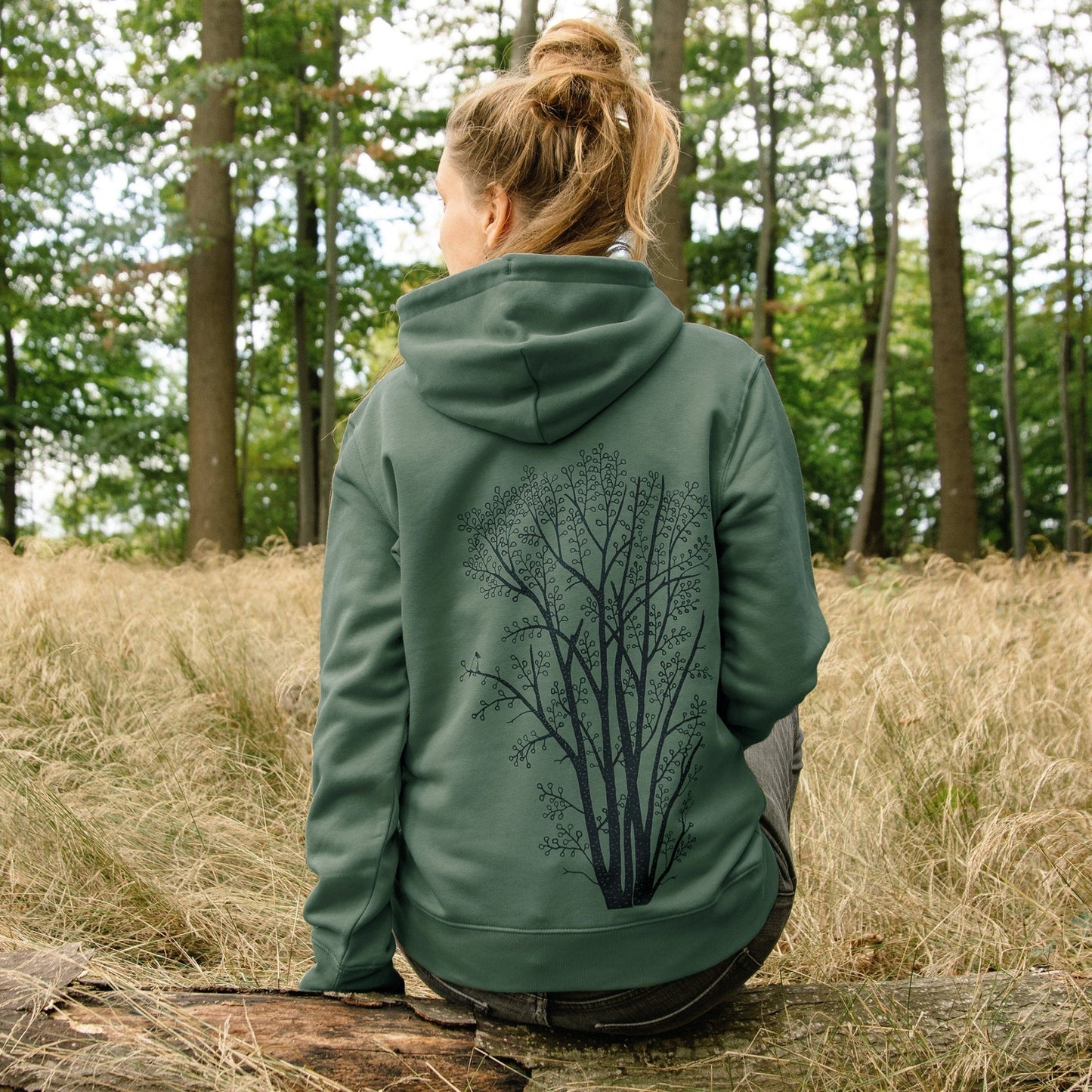 Hoodie mit Baum Print als gemütliche Herbstmode für Damen. Eine Frau sitzt auf einem Baumstamm im Wald und präsentiert das nachhaltige Pflanzen Motiv aus Bio-Baumwolle.
