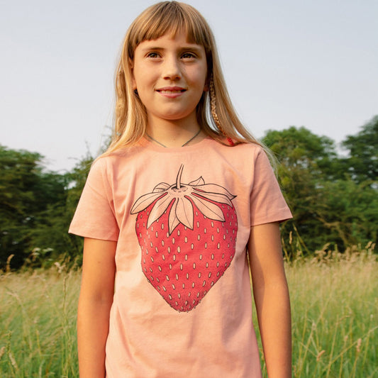 Kinder Erdbeer T-Shirt für Mädchen mit großem Früchte-Motiv. Ein kleines Mädchen präsentiert die Vorderansicht des handbedruckten Bio-Shirts mit der großen Erdbeere von vorne.
