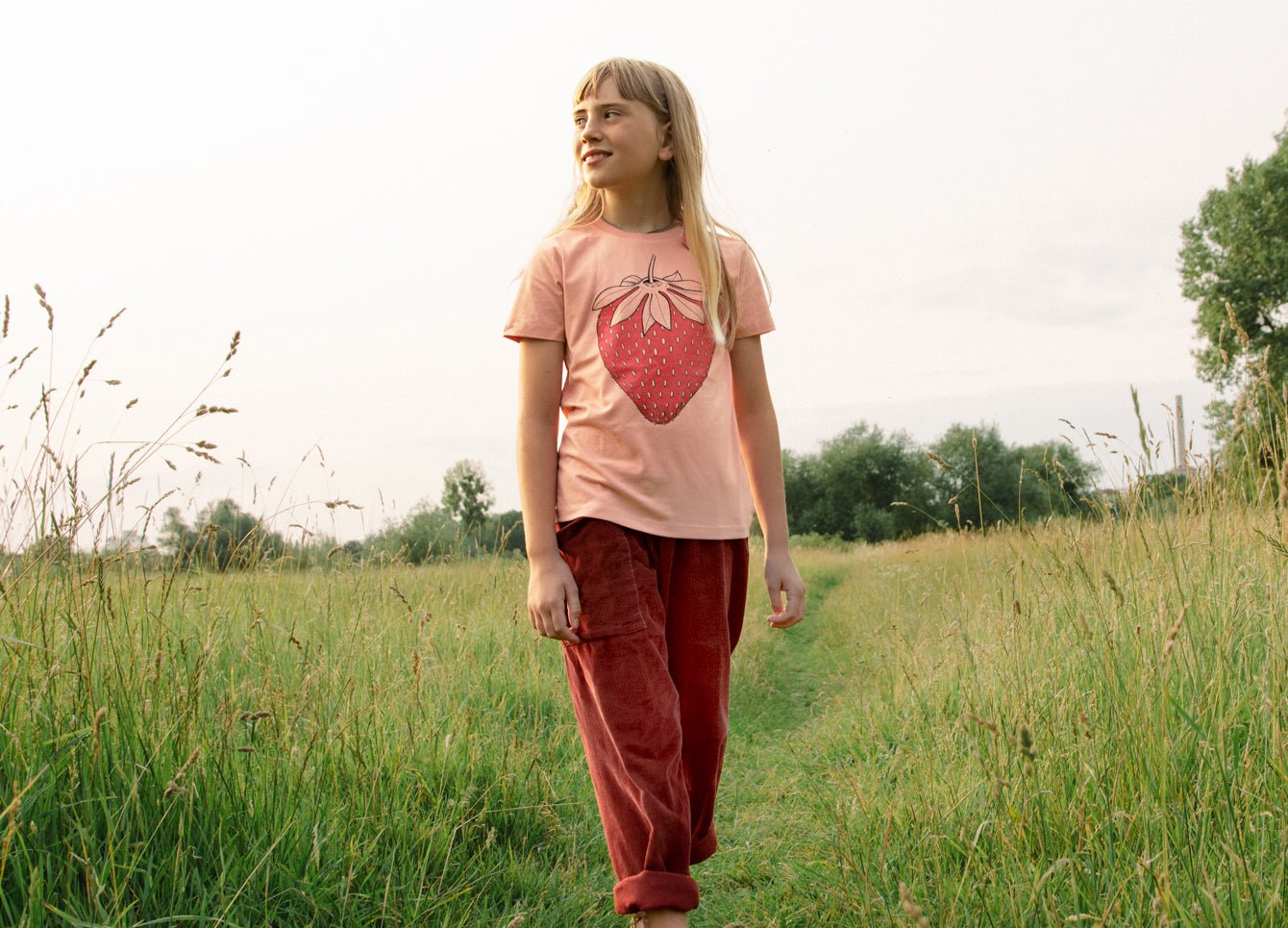 Kinder Erdbeer Tshirt aus nachhaltiger Bio-Baumwolle für den Sommer in rosa. Ein Outdoor-Lifestyle-Bild zeigt ein kleines Mädchen in Ganzkörperansicht in der Natur mit dem bunt bedruckten Oberteil.
