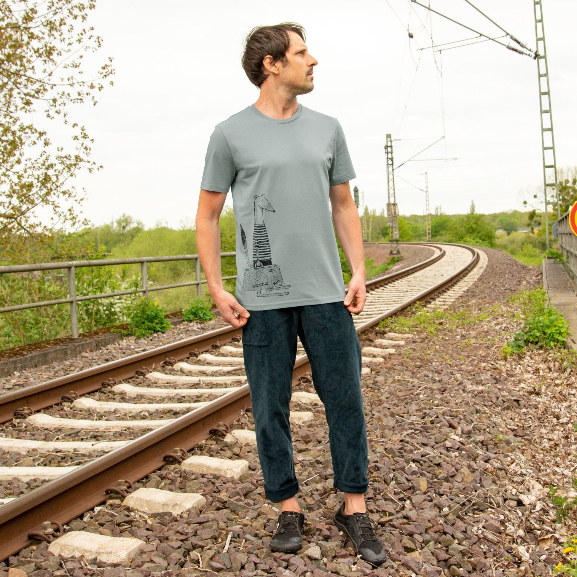 T-Shirt Wiesel für Männer im bequemen Alltags- und Reise-Look. Ein Outdoor-Lifestyle-Bild zeigt einen Mann in Ganzkörperansicht mit dem handbedruckten Naturmode-Oberteil aus Bio-Baumwolle.

