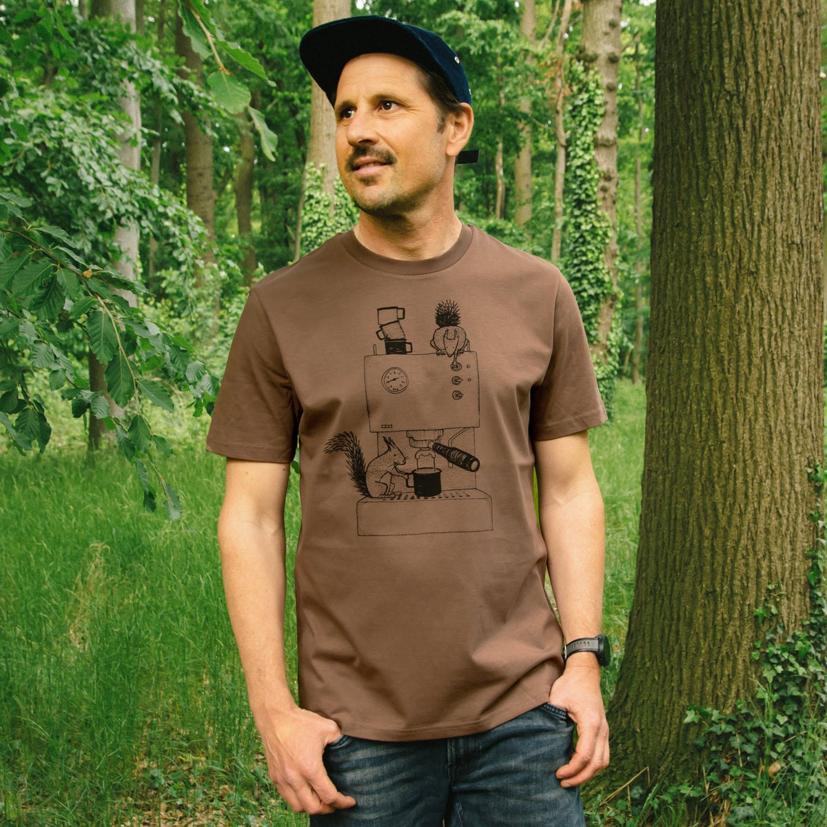 Kaffee T-Shirt für Herren im dunklen Farbton Mocca. Ein Mann steht im Wald und präsentiert das kreative Eichhörnchen-Motiv als hochwertigen Frontprint auf der Vorderseite des Bio-Shirts.