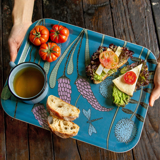 Schwedisches Design Holztablett beim Servieren von Frühstück im Garten. Eine Person trägt das dunkelblaue Serviertablett mit floralem Muster, beladen mit einem frischen Baguette, Tomaten und unserer handillustrierten Cmig Emaille-Tasse