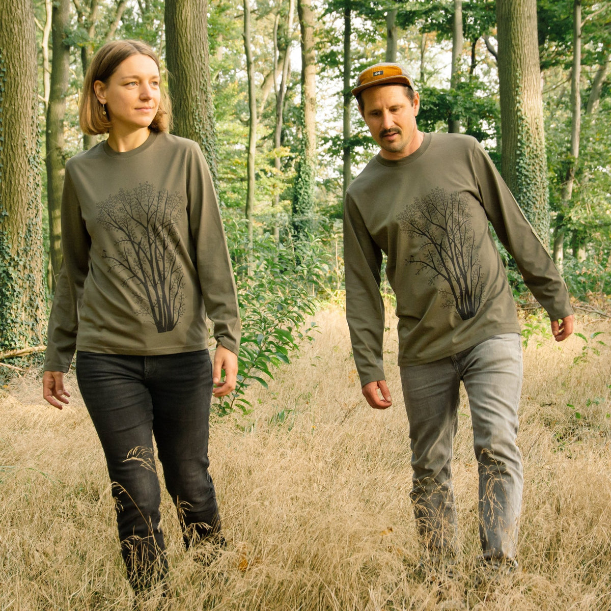 Tshirt Langarm Baum Print als sportlicher Partnerlook in Khaki. Eine Frau und ein Mann stehen in Ganzkörperansicht im herbstlichen Wald und tragen das handbedruckte Bio-Oberteil.
