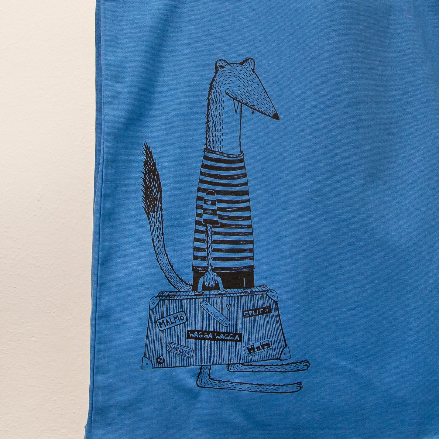 Witziges Wiesel-Motiv mit Reisekoffer als Siebdruck auf einer blauen Stofftasche. Detailaufnahme der kunstvollen Urlaubs-Illustration, gefertigt mit umweltfreundlichen, wasserbasierten Textildruckfarben.
