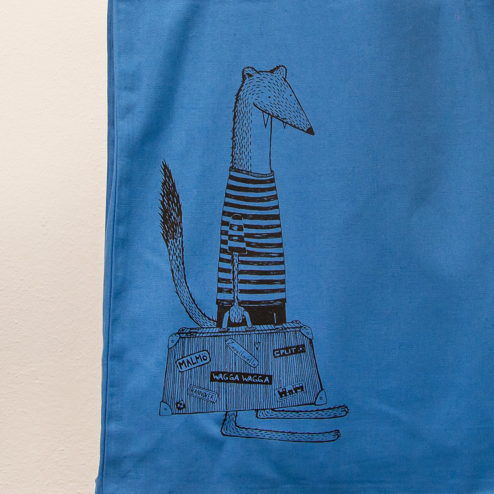 Witziges Wiesel-Motiv mit Reisekoffer als Siebdruck auf einer blauen Stofftasche. Detailaufnahme der kunstvollen Urlaubs-Illustration, gefertigt mit umweltfreundlichen, wasserbasierten Textildruckfarben.
