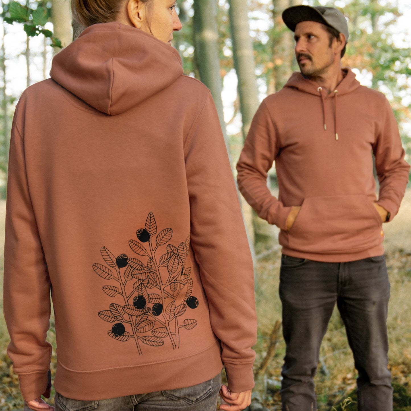 Blaubeere Hoodie als nachhaltige Naturmode für Damen und Herren. Ein herbstliches Outdoor-Lifestyle-Bild im Wald zeigt ein Pärchen im braunen Partnerlook-Kapuzenpulli.

