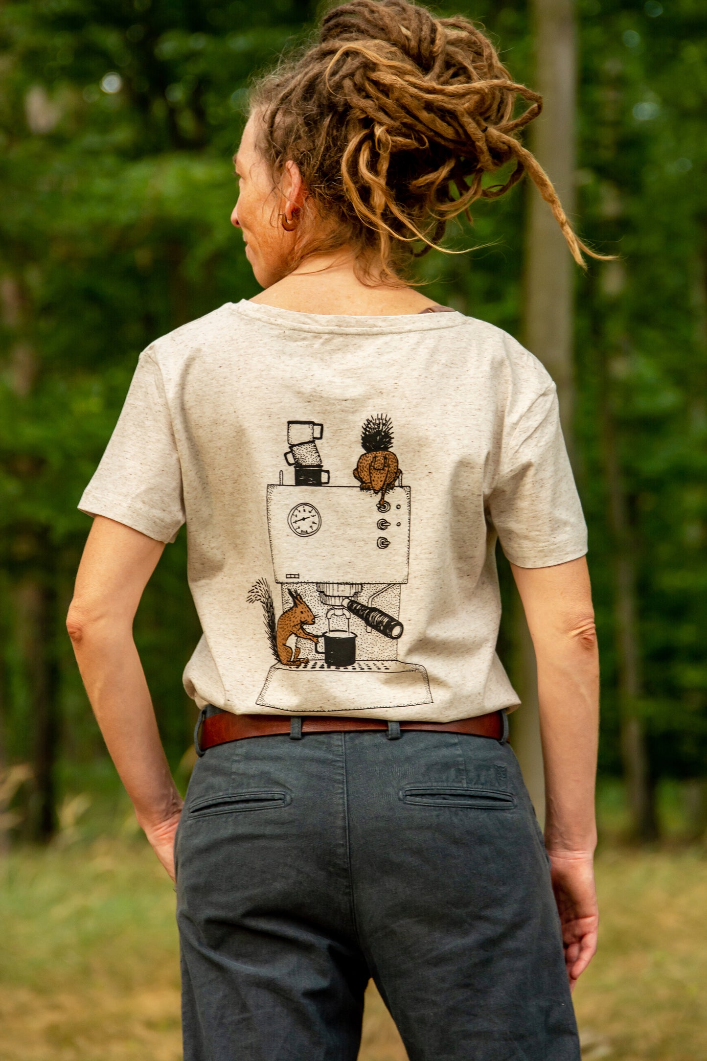 Kaffee T-Shirt für Damen, getragen von einer Frau, die im Wald steht. Das locker in die Hose eingesteckte Shirt zeigt den detailreichen Backprint mit dem Eichhörnchen an der Espressomaschine.