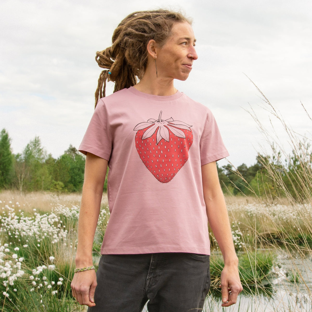 Erdbeer T-Shirt für Damen im lockeren, kurzen Schnitt. Eine Frau steht in der Natur und präsentiert die Vorderansicht des naturfarbenen Bio-Shirts mit der großen Früchte-Illustration.
