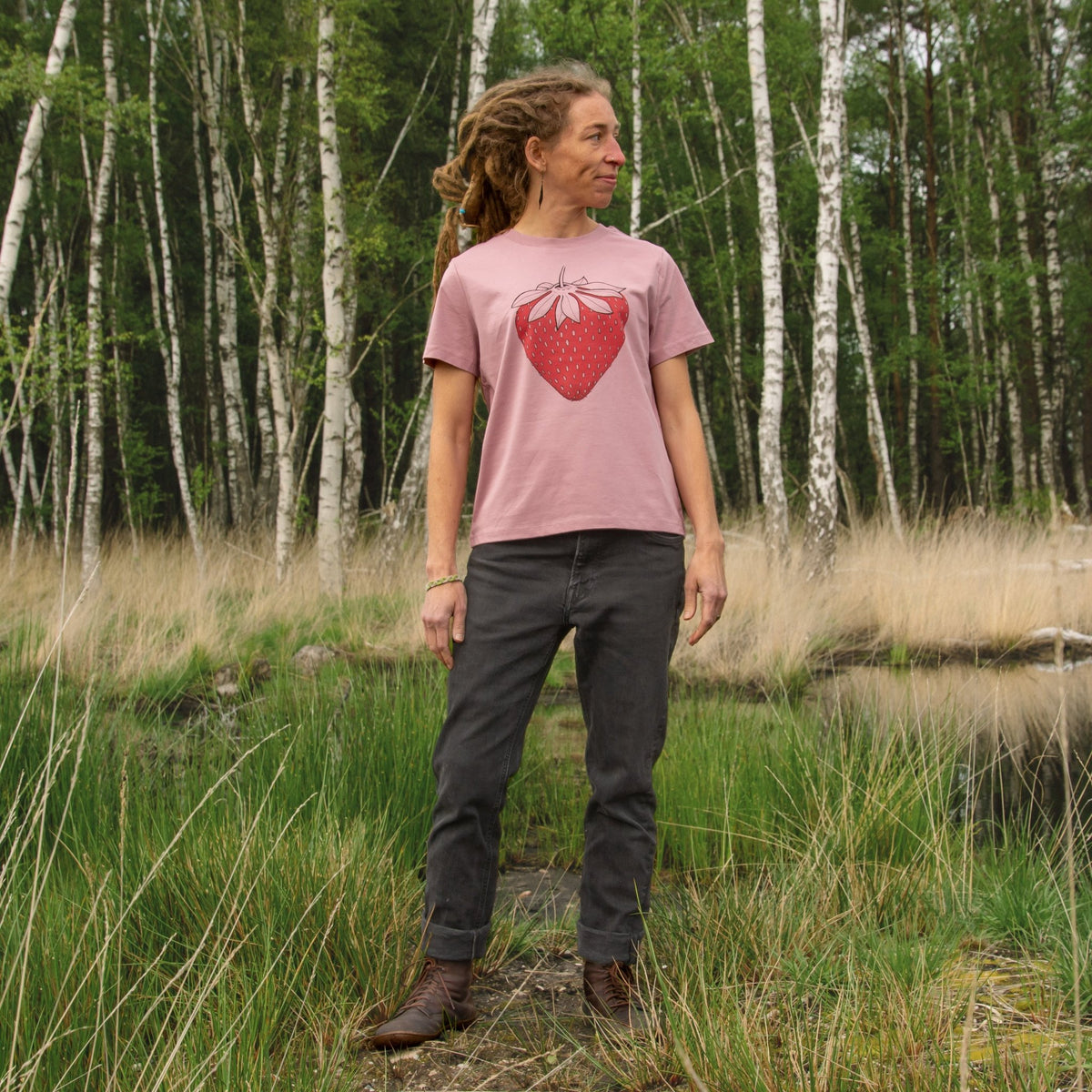 Erdbeer Tshirt für Damen getragen im lässigen Sommer-Look. Ein Outdoor-Lifestyle-Bild zeigt eine Frau in Ganzkörperansicht in der Natur mit dem handbedruckten botanischen Oberteil.
