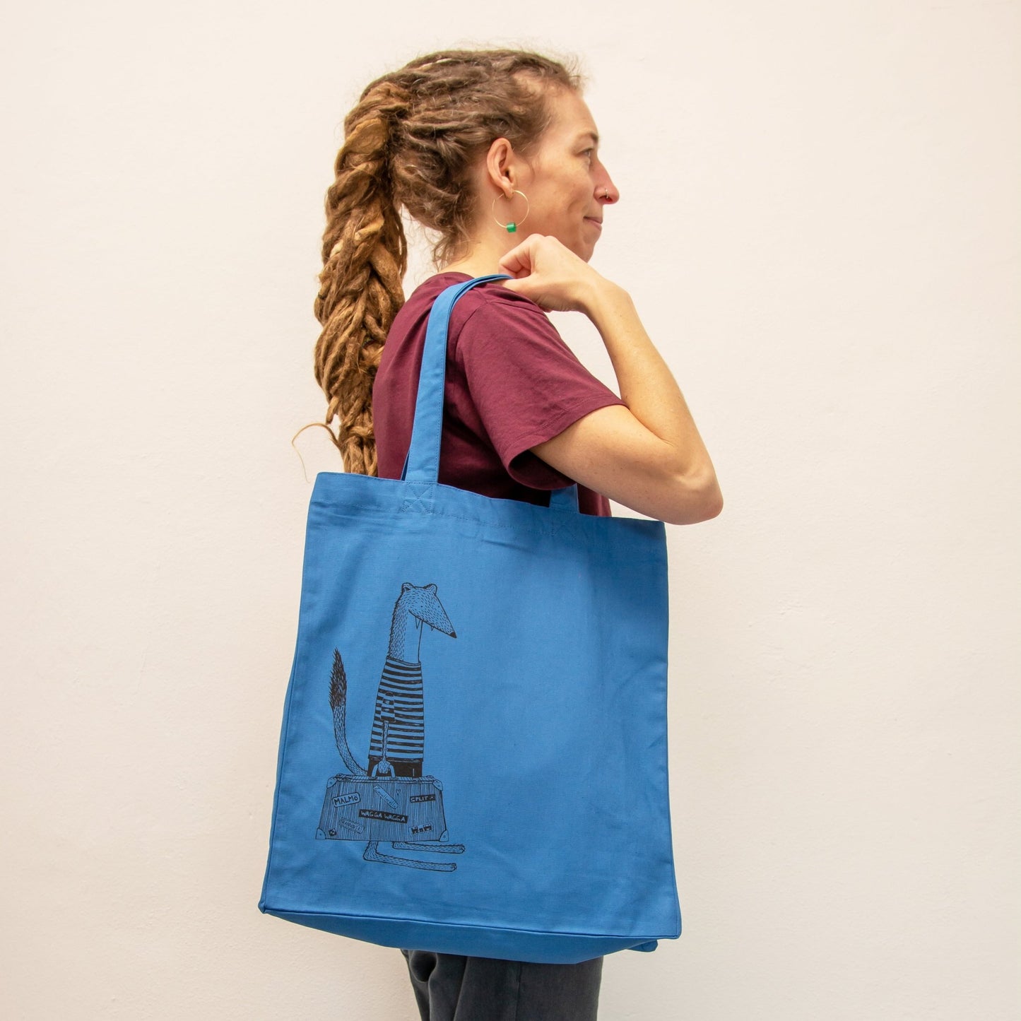 Wiesel Jutebeutel in Mindful Blue getragen von einer Frau. Der rauchblaue Shopper mit Tiermotiv zeigt sich als praktischer Alltagsbegleiter und kreatives Geschenk für Weltenbummlerinnen.
