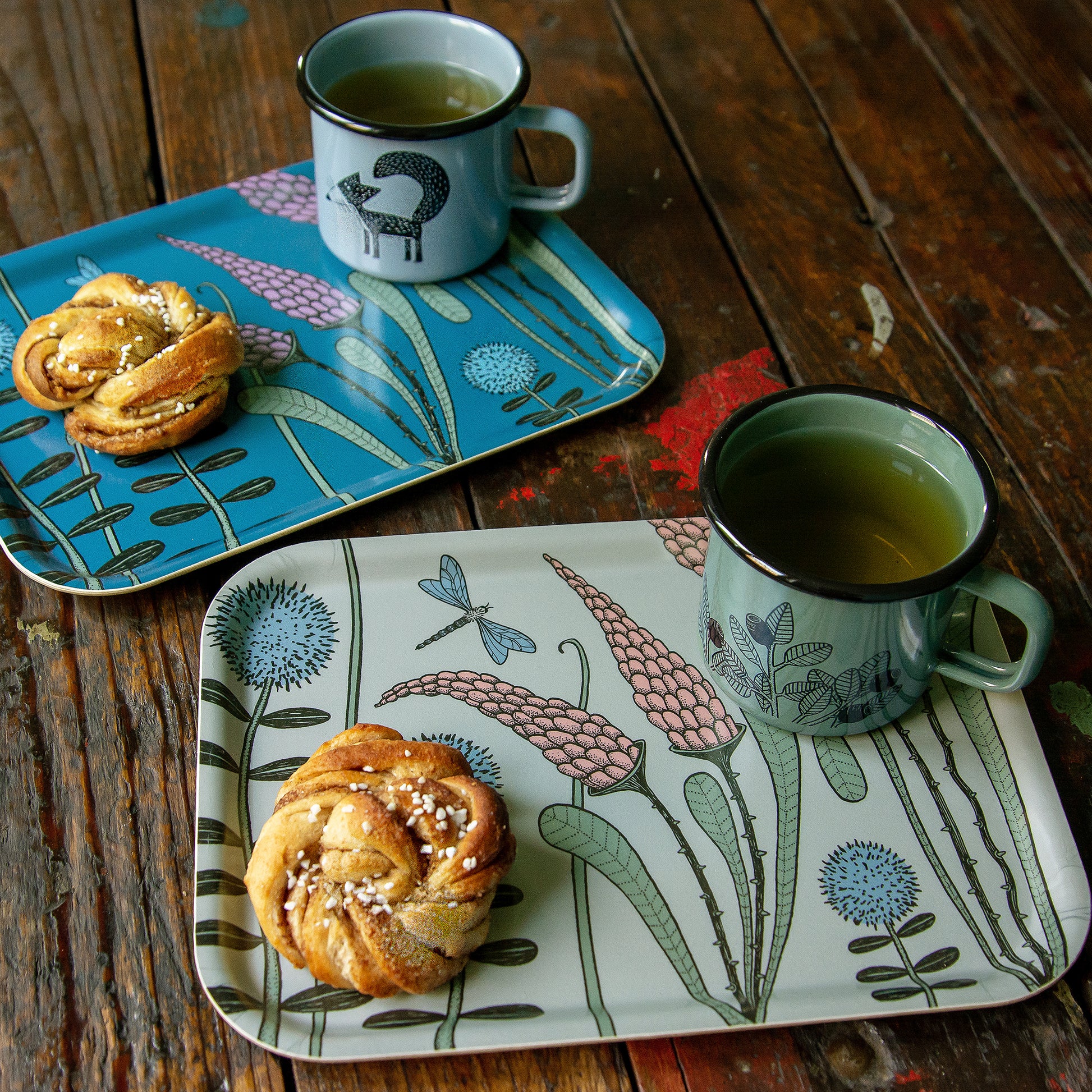 Deko-Tabletts aus Holz im schwedischen Design mit Cmig Emaille-Tassen. Salbeigrünes Fika-Tablett mit Blaubeer-Tasse und Zimtschnecke, im Hintergrund dekoriert mit dem dunkelblauen Waldwiese-Tablett und der hellblauen Fuchs-Emailletasse.