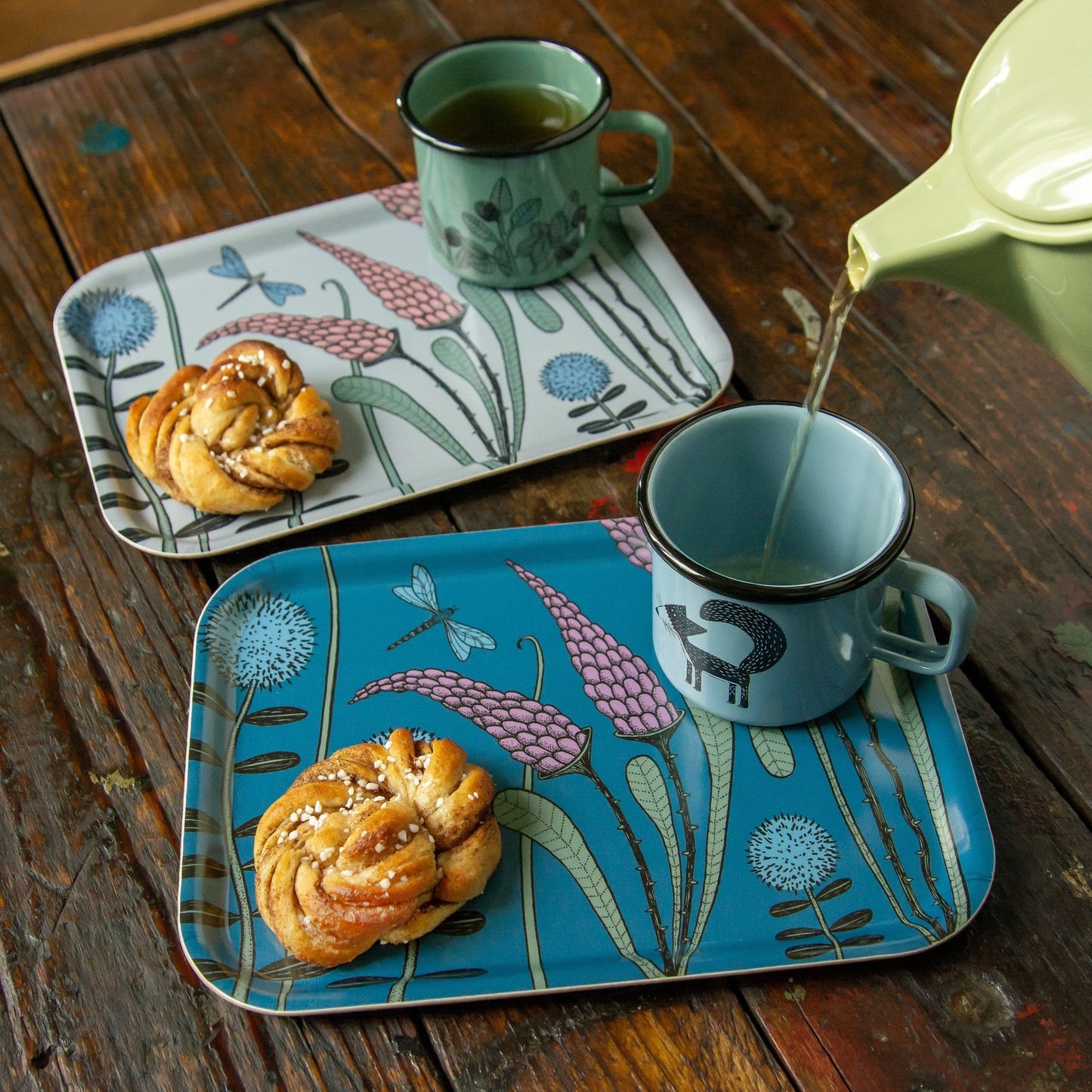 Kaffee-Tablett im Scandi-Style aus Birkenfurnier mit schwedischer Zimtschnecke. Kleines dunkelblaues Deko-Tablett mit Wiesenblumen-Design, gemütlich gedeckt mit einer Kaffeetasse und einer frischen Zimtschnecke auf dem Tisch.