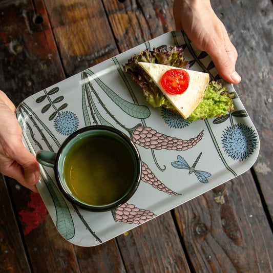 Schwedisches Design Holztablett beim Servieren von Frühstück im Garten. Eine Person trägt das salbeigrüne Serviertablett mit floralem Muster, beladen mit einer Tasse Tee und einem frischen, belegten Brot.