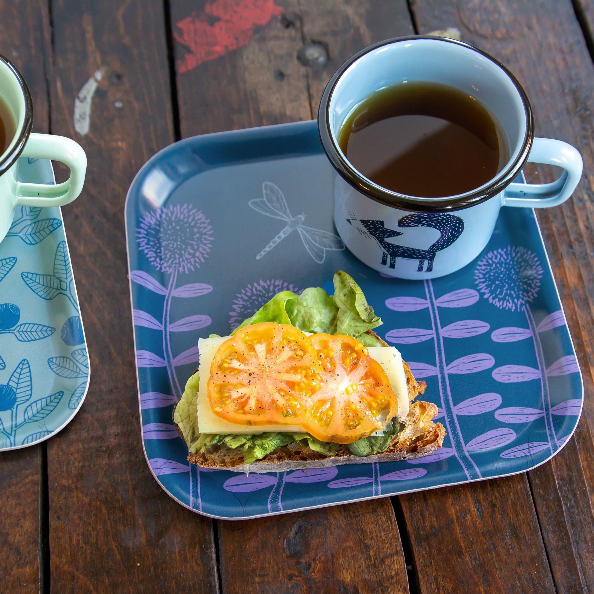 Kleiner Scandi-Style Frühstücksteller aus Holz mit Cmig Fuchstasse. Blau-lilanes Serviertablett mit Wiesenblumen-Motiv, angerichtet mit einem belegten Baguette und unserer beliebten hellblauen Emailletasse mit Fuchs-Motiv.