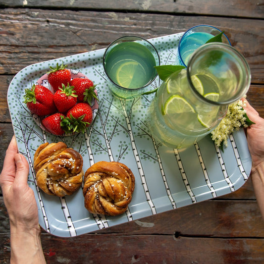Schwedisches Design Holztablett für die gemütliche Fika im Garten. Hellblaues Serviertablett mit Birken-Muster, sommerlich gedeckt mit frischen Zimtschnecken, roten Erdbeeren und erfrischendem Holundersaft.