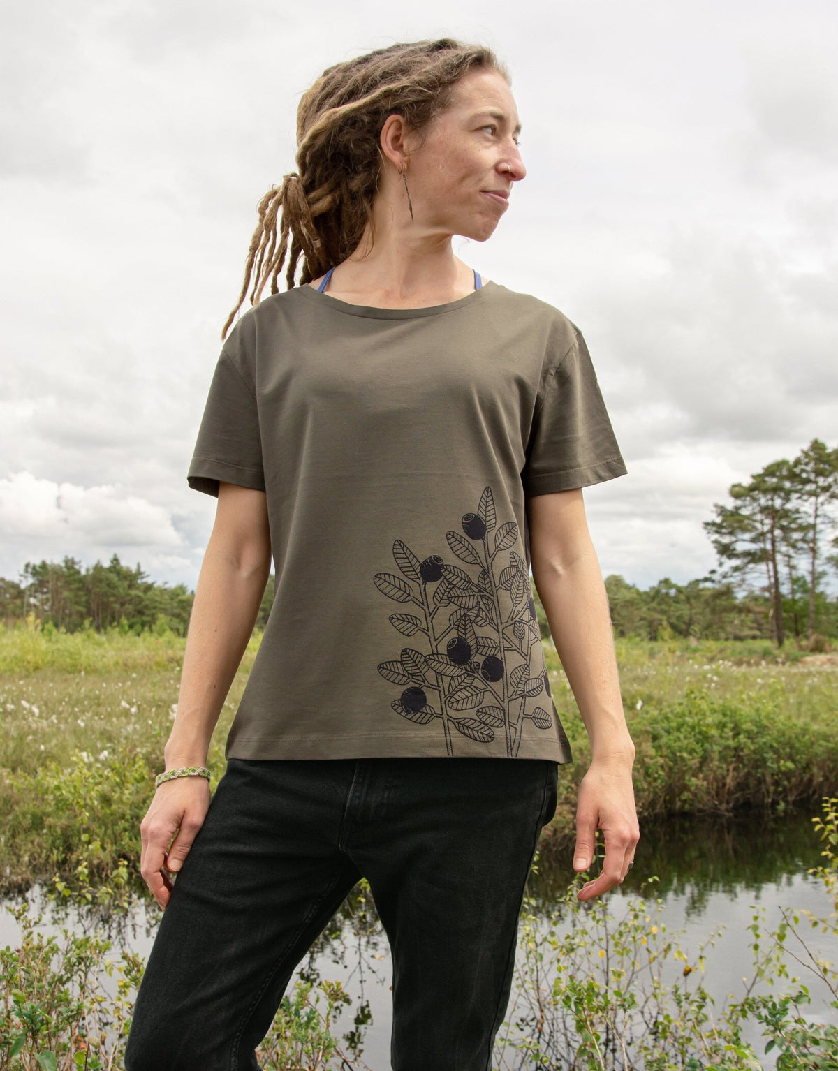 Blaubeere Tshirt für Damen aus dickerem Bio-Stoff (180g/m²). Ein herbstliches Outdoor-Lifestyle-Bild zeigt eine Frau in Ganzkörperansicht in der Natur mit dem handbedruckten oliv-khaki Oberteil.
