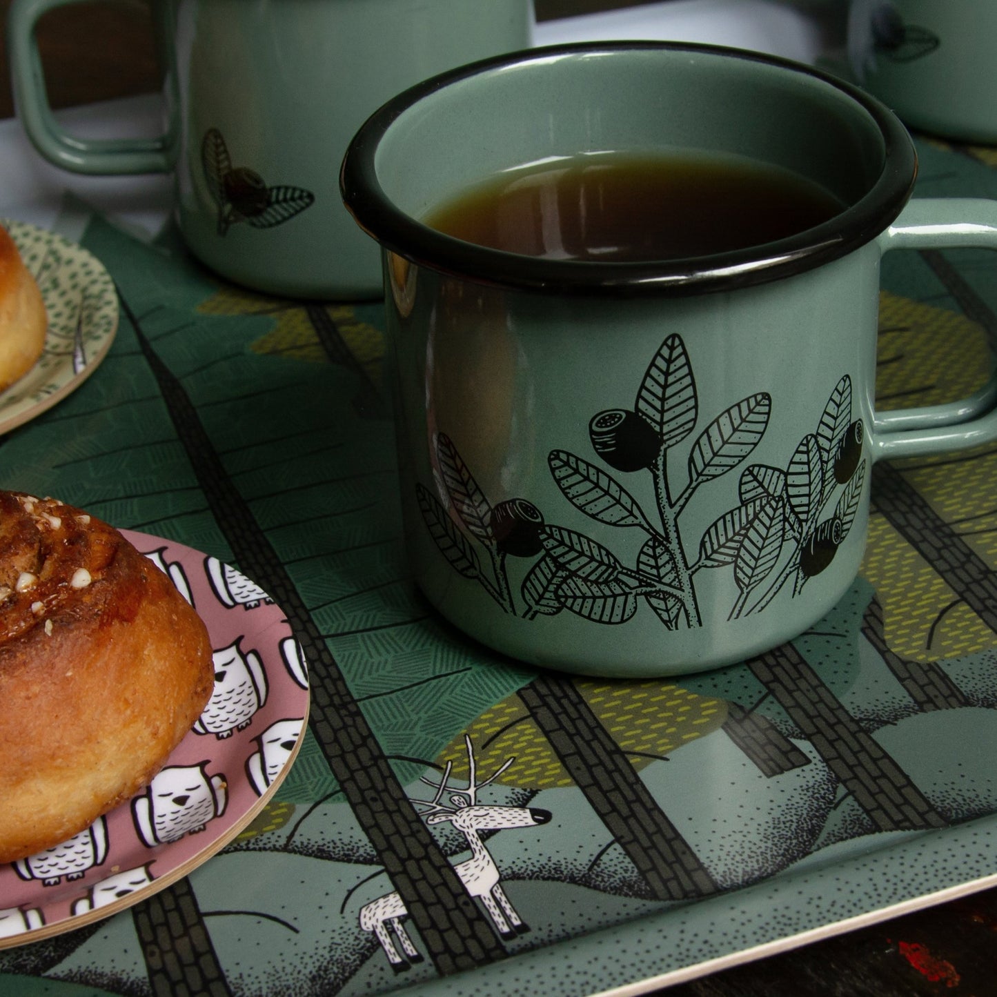 Schwedisches Design Holztablett mit Tannen-Muster und waldgrüner Emailletasse. Detailansicht des großen Serviertabletts mit kleinem Rentier im Wald, perfekt kombiniert mit unserer handillustrierten Cmig Blaubeer-Tasse für Outdoorfans.