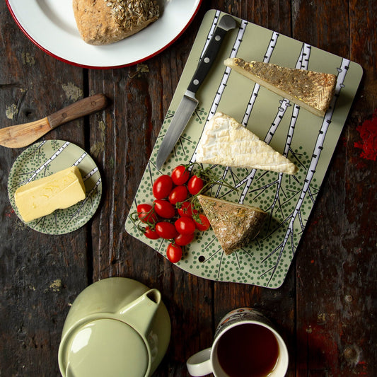 Schwedisches Design Frühstücksbrettchen aus Holz reichlich gedeckt. Grünes Servierbrett mit Pflanzen-Muster, angerichtet mit einem Messer, frischem Käse und Tomaten inmitten von gemütlichen Frühstückssachen.